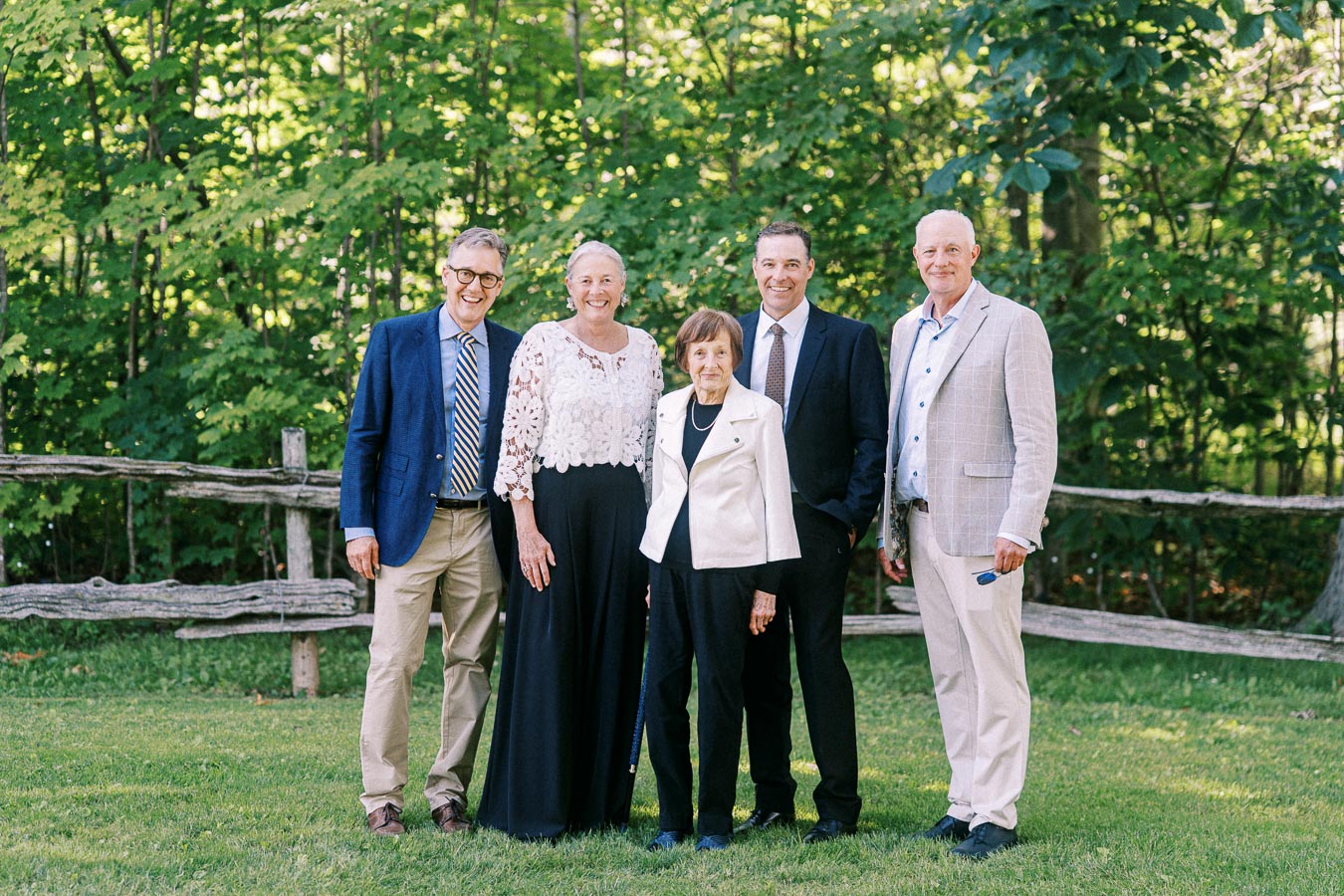 Five adult wedding guests pose outdoors on a grassy private estate near Chatham-Kent, Ontario, with a rustic wooden fence and green trees behind them.