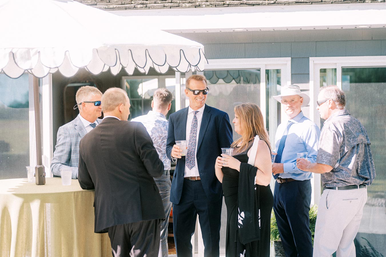 Wedding guests in formal attire socialize with drinks outside a private estate near Chatham-Kent, Ontario, beside a white tent canopy.