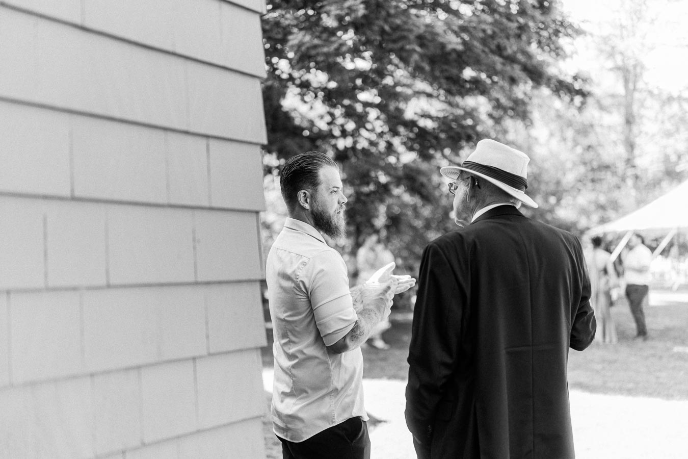 Black and white photo of two men talking beside a barn wall during an outdoor tented wedding on a private farm estate near Chatham-Kent, Ontario.