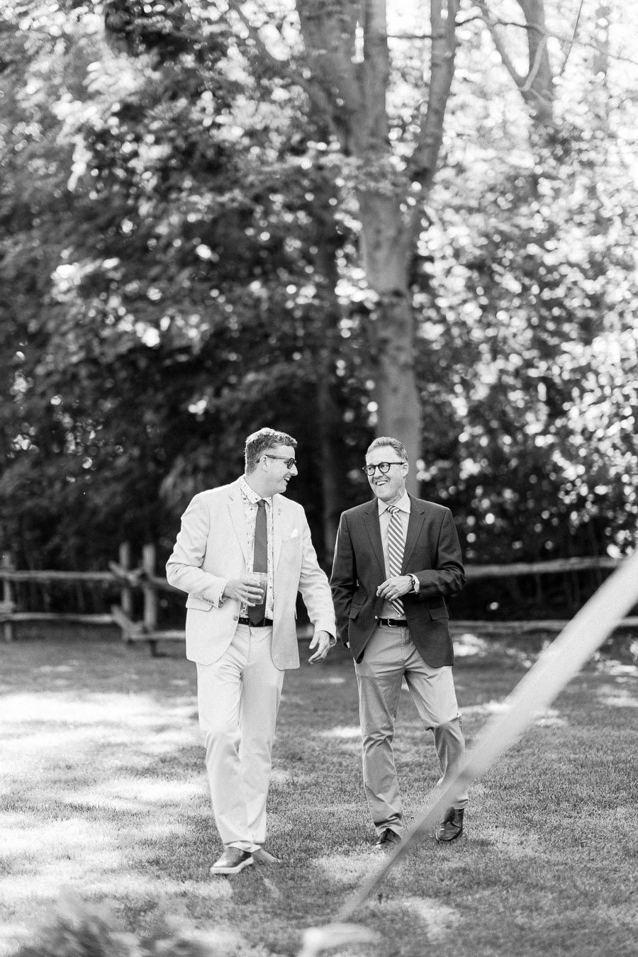 Two male wedding guests in suits walk and talk on a grassy estate lawn near Chatham-Kent, Ontario, in black and white.