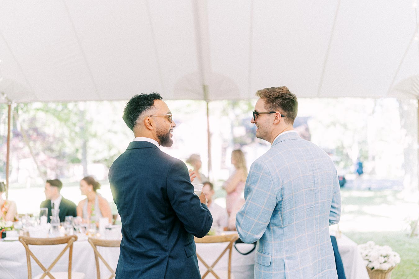 Two men in suits talking during a tented outdoor wedding reception on a private estate near Chatham-Kent, Ontario, Canada.
