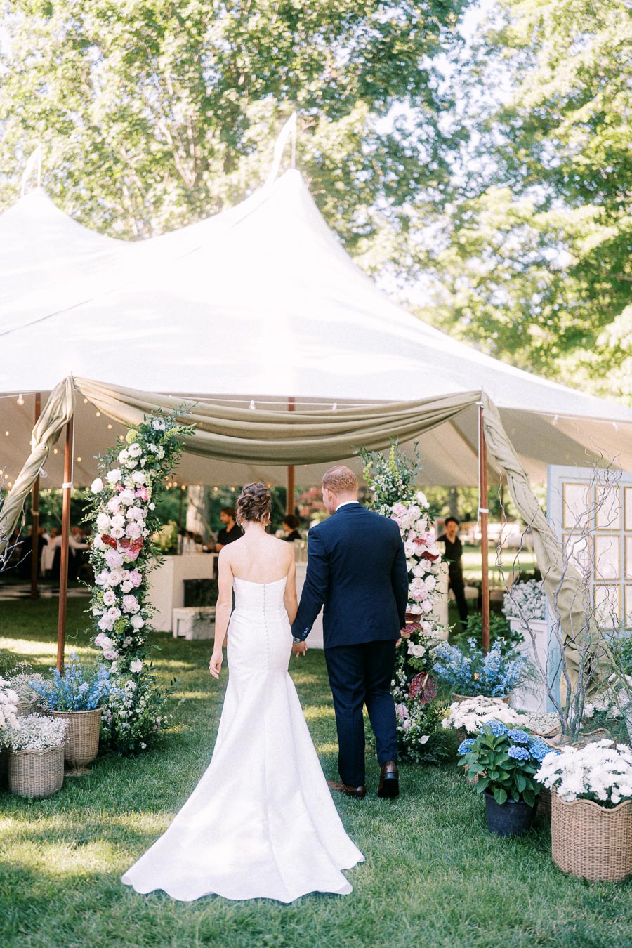 Bride and groom walking hand in hand into a white wedding tent decorated with floral arches on a private farm estate near Chatham-Kent, Ontario.