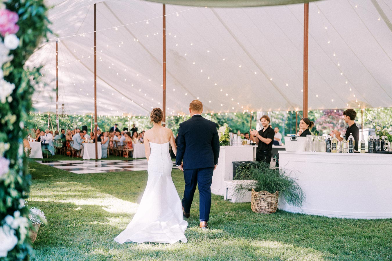 Bride and groom enter a white tented wedding reception on a grassy private estate near Chatham-Kent, Ontario, with guests, bar, and string lights.