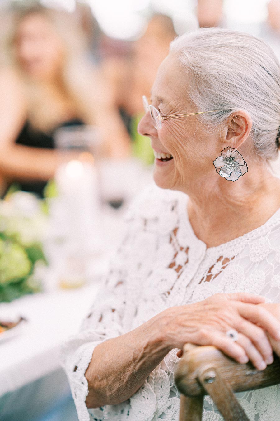 Smiling elderly woman in a white lace blouse and floral earrings seated at an outdoor wedding dinner near Chatham-Kent, Ontario, Canada.