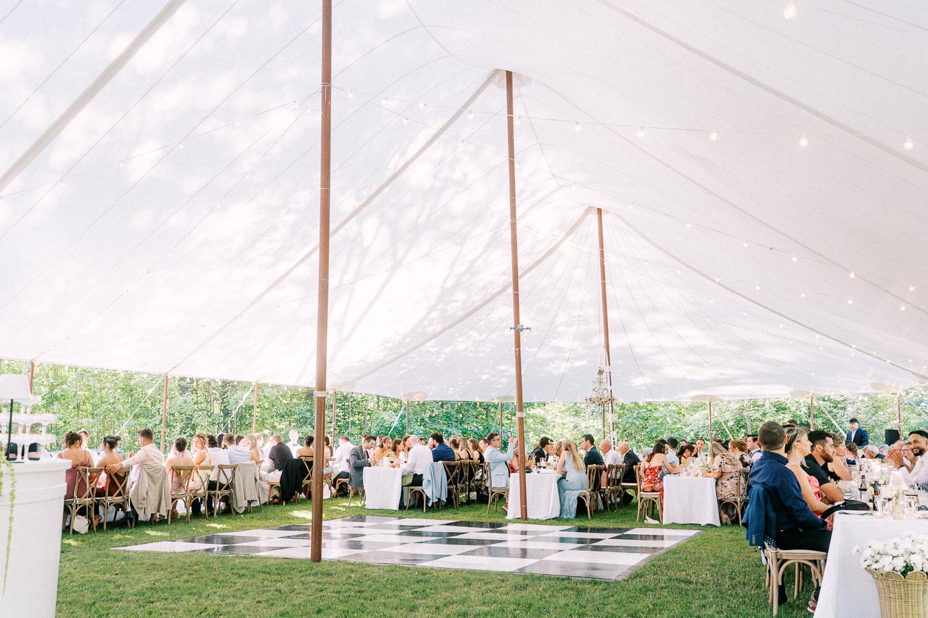 Guests dining under a white wedding tent with string lights, long tables, wooden chairs, and a checkered dance floor near Chatham-Kent, Ontario.