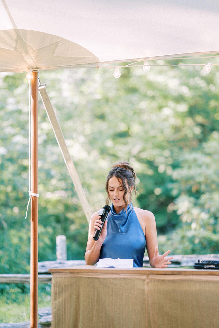 Woman in a blue dress speaks into a microphone under a wedding tent during an outdoor private estate reception near Chatham-Kent, Ontario.