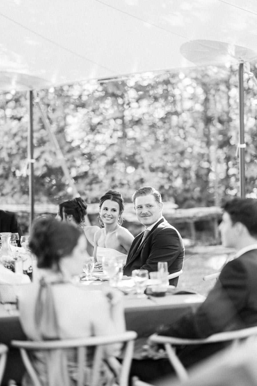 Black and white photo of a bride and groom seated with guests at an outdoor tented wedding dinner on a private farm estate near Chatham-Kent, Ontario.
