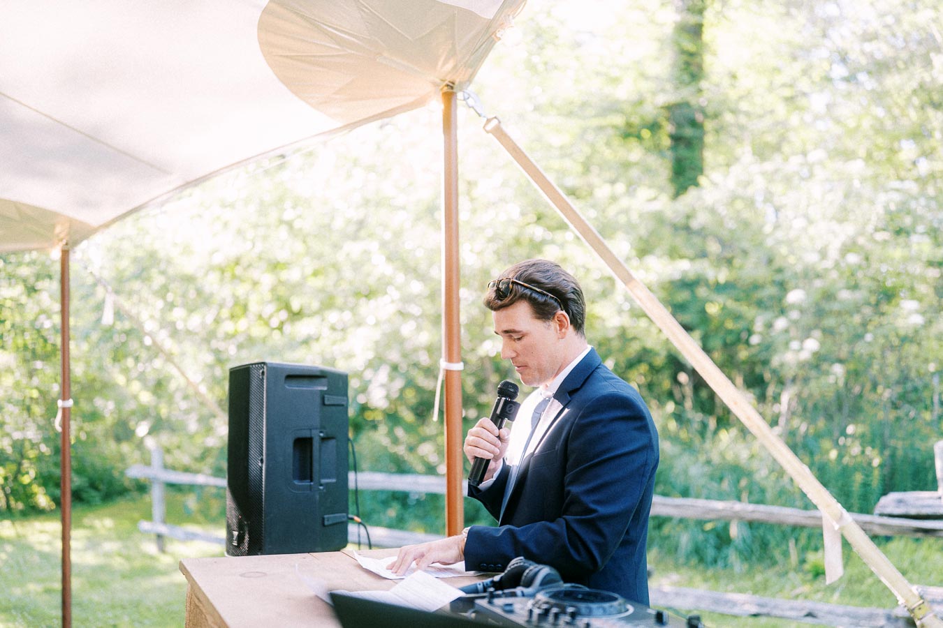 Man in a navy suit speaking into a microphone at a tented outdoor wedding reception on a private farm estate near Chatham-Kent, Ontario, Canada.