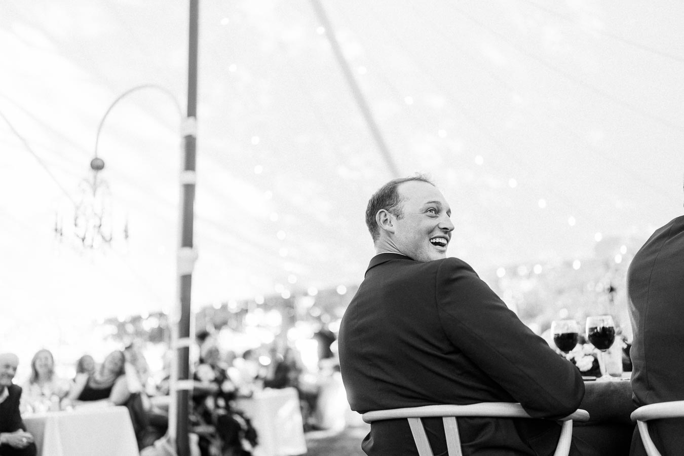 Black and white photo of a smiling man in a suit seated under a wedding tent during an outdoor farm reception near Chatham-Kent, Ontario.