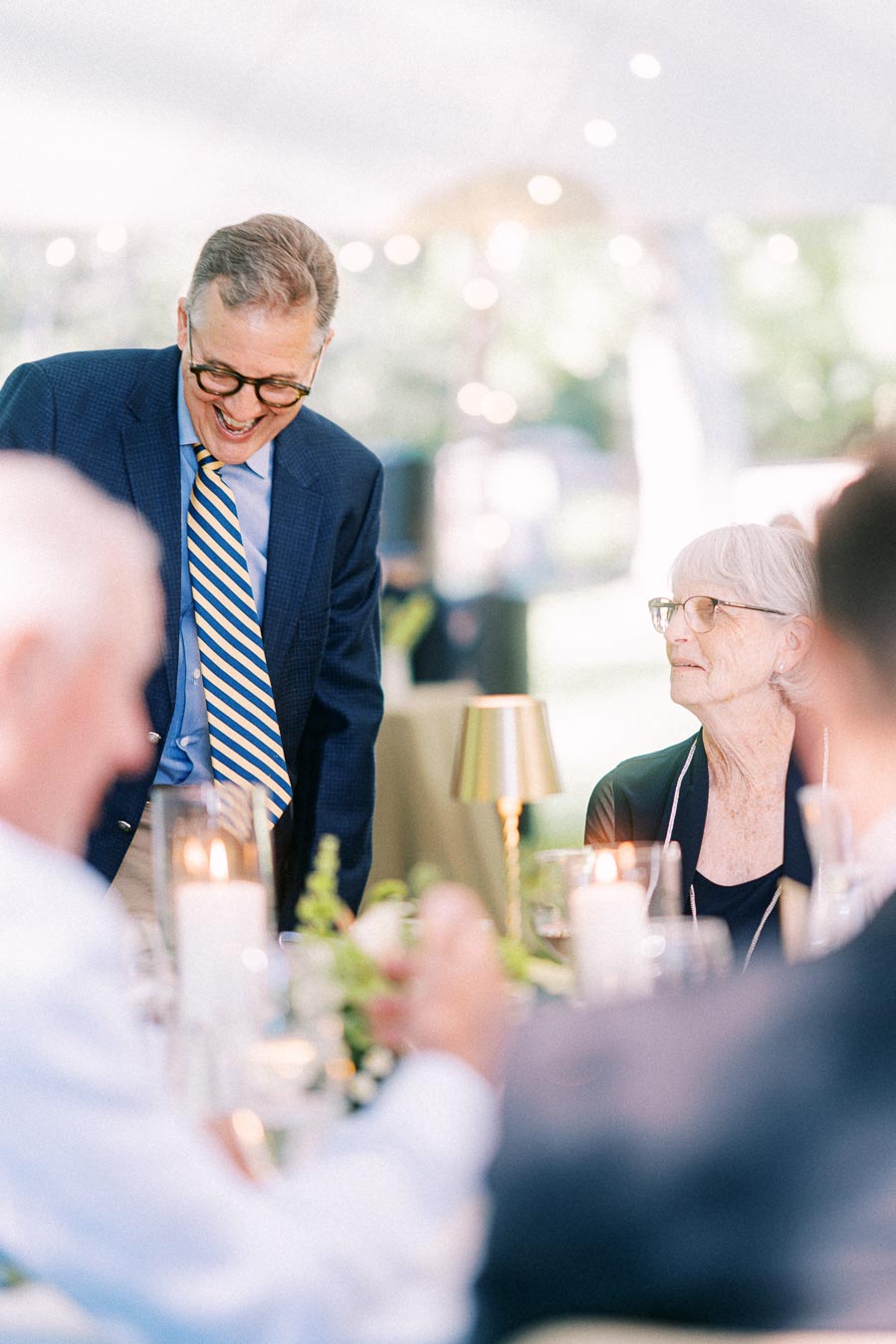 Smiling man in blue suit speaks with an elderly woman during a candlelit tented wedding dinner near Chatham-Kent, Ontario, Canada.