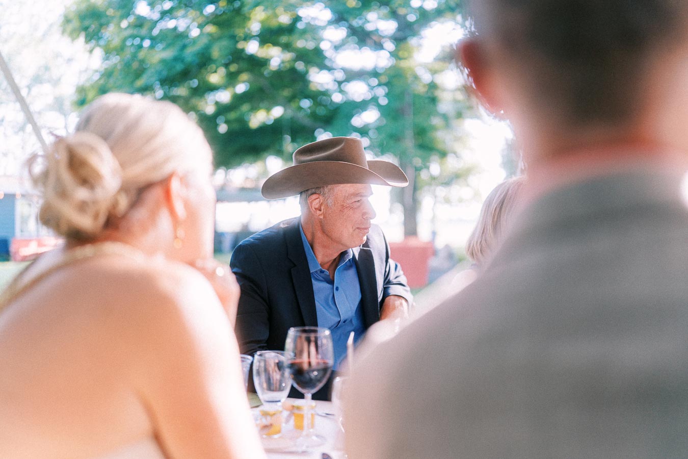 Wedding guests seated at an outdoor farm reception table near Chatham-Kent, Ontario, with a man in a cowboy hat in focus.