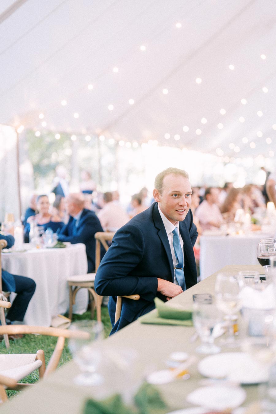 Wedding guest in a suit seated at a tented outdoor reception near Chatham-Kent, Ontario, with tables, glassware, and string lights.