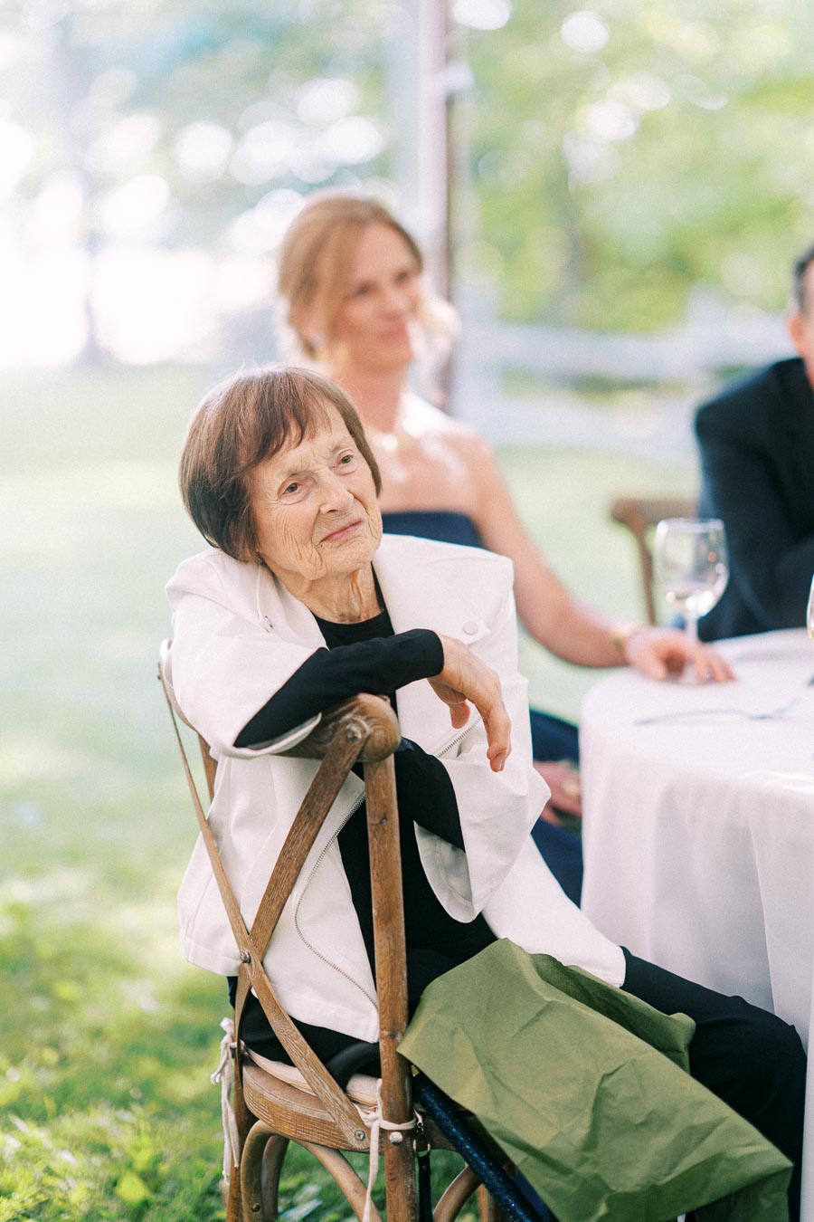 Elderly woman seated at an outdoor tented wedding reception near Chatham-Kent, Ontario, with guests and table setting in soft daylight.