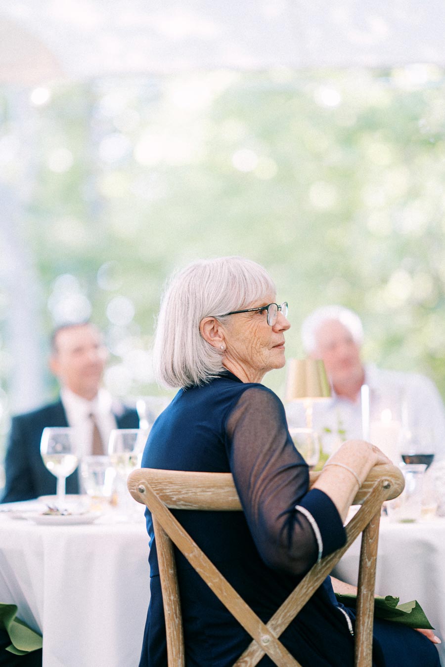 Older woman with glasses seated at a tented outdoor wedding reception near Chatham-Kent, Ontario, with guests and table settings behind her.