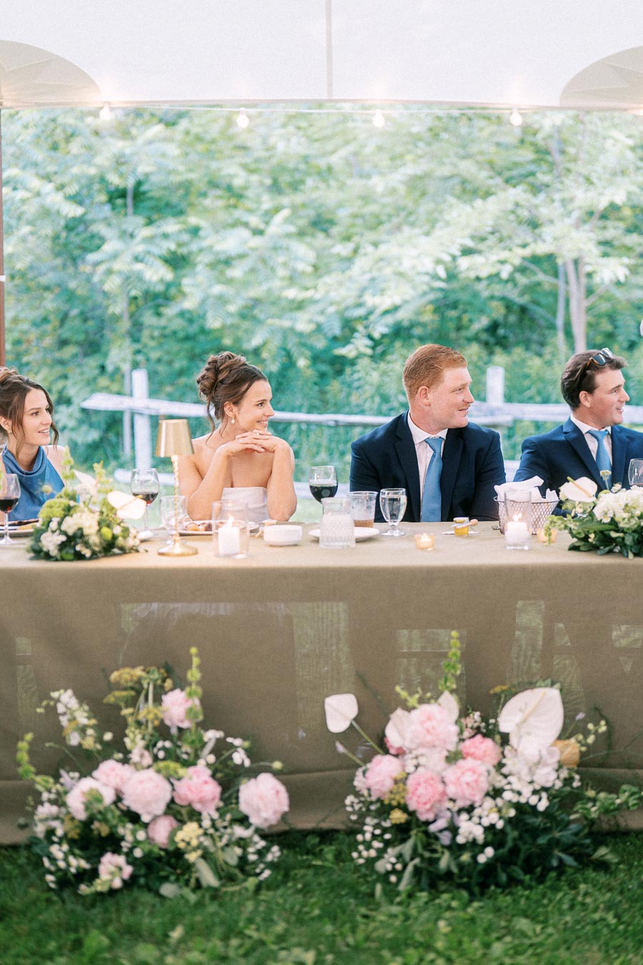Bride and groom seated with wedding guests at a decorated head table under a tent during an outdoor private estate wedding near Chatham-Kent, Ontario, Canada.