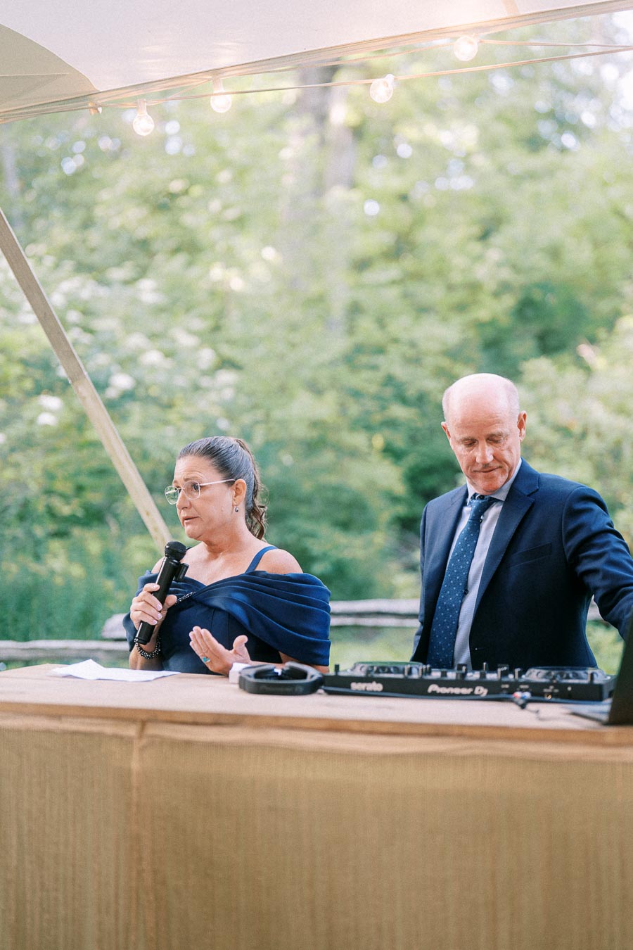 Woman speaking into a microphone beside a suited DJ at an outdoor tented wedding reception near Chatham-Kent, Ontario, Canada.