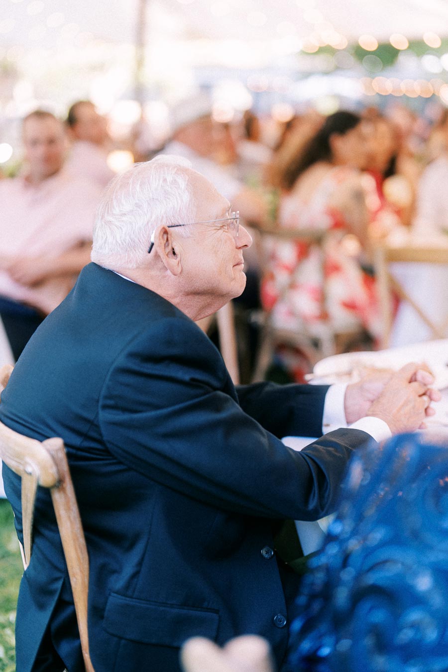 Elderly man in a dark suit seated at a tented outdoor wedding reception near Chatham-Kent, Ontario, with blurred guests and warm lights.