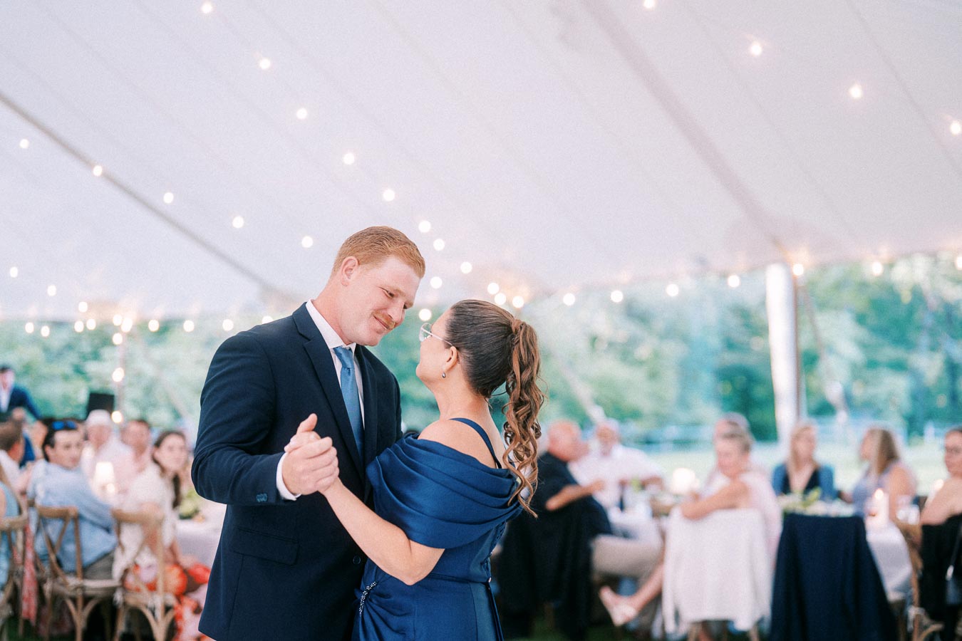Man in a navy suit dances with a woman in a blue dress under a white wedding tent with string lights and seated guests in Chatham-Kent, Ontario.