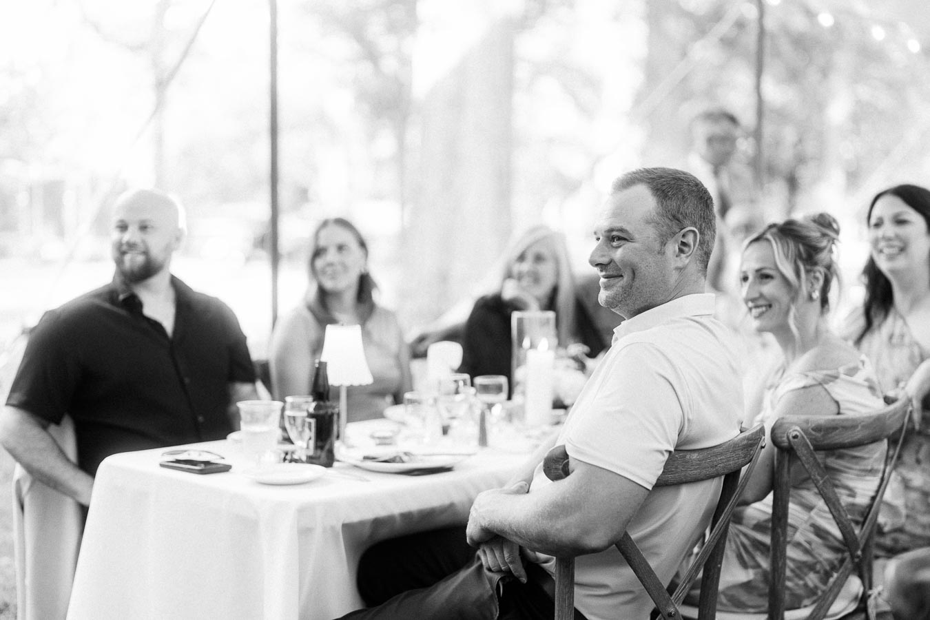 Black and white photo of wedding guests smiling at a round dinner table under an outdoor tent near Chatham-Kent, Ontario, Canada.