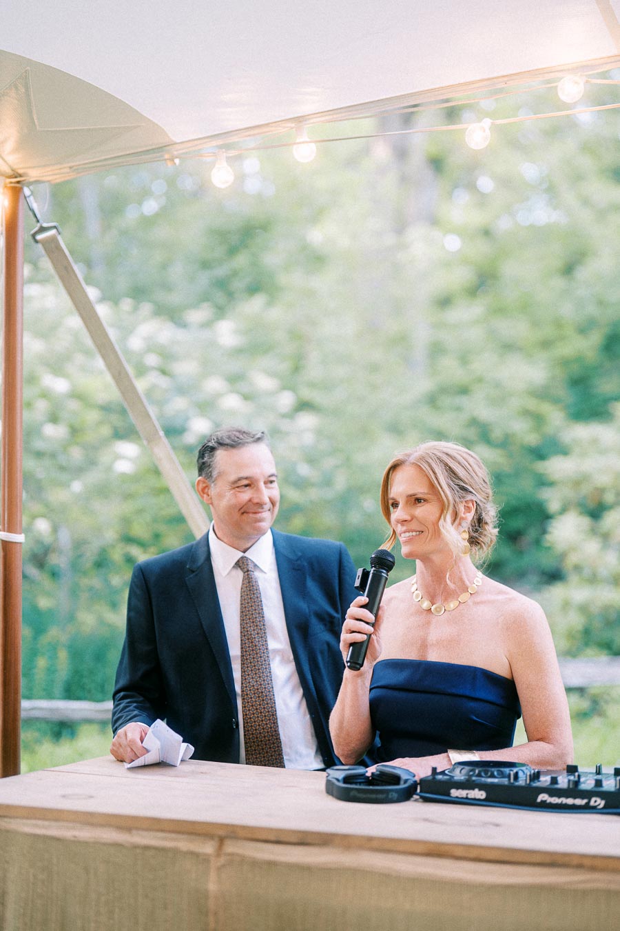 Woman speaking into a microphone beside a man under a wedding tent with string lights, DJ equipment, and greenery near Chatham-Kent, Ontario.