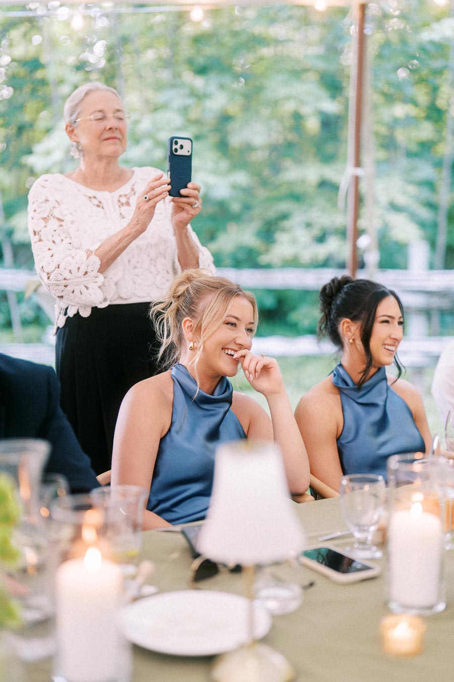 Wedding guests laughing at a tented outdoor reception near Chatham-Kent, Ontario, with candles, tableware, and a woman taking photos on a phone.