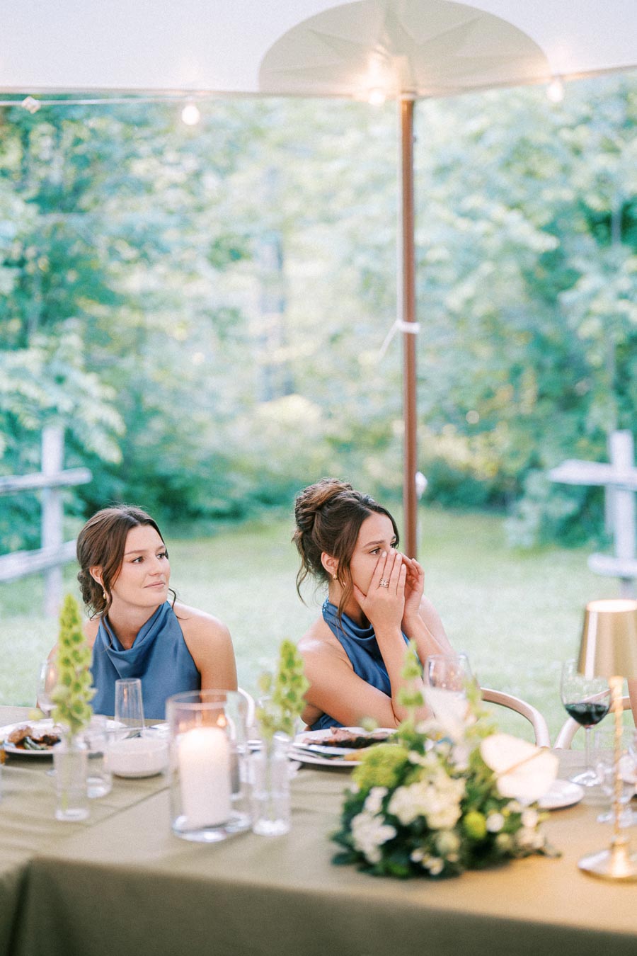 Two bridesmaids sit at an outdoor farm wedding reception table under a tent on a private estate near Chatham-Kent, Ontario, with candles, flowers, wine glasses, string lights, and green countryside in the background.