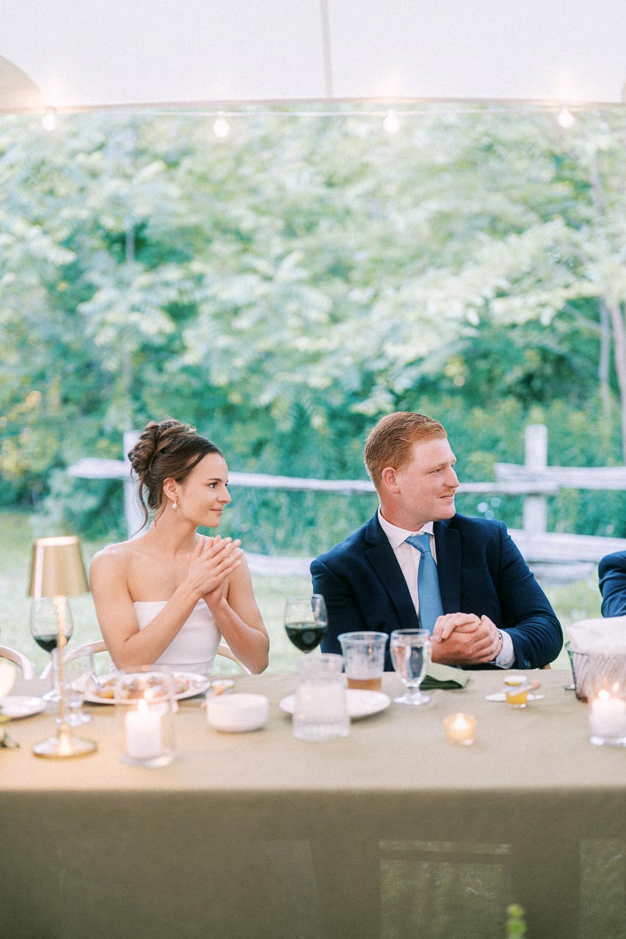 Bride and groom seated at a tented outdoor wedding dinner on a private farm estate near Chatham-Kent, Ontario, with candles, wine glasses, and greenery.