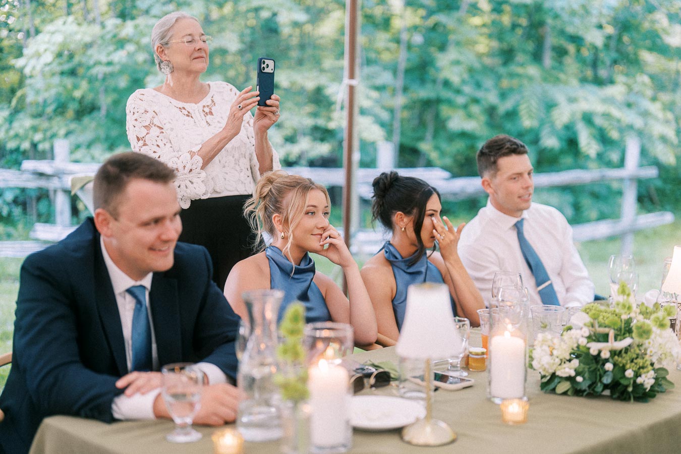 Wedding guests at a tented outdoor dinner on a private estate near Chatham-Kent, Ontario, with candles, flowers, and a woman recording on a smartphone.