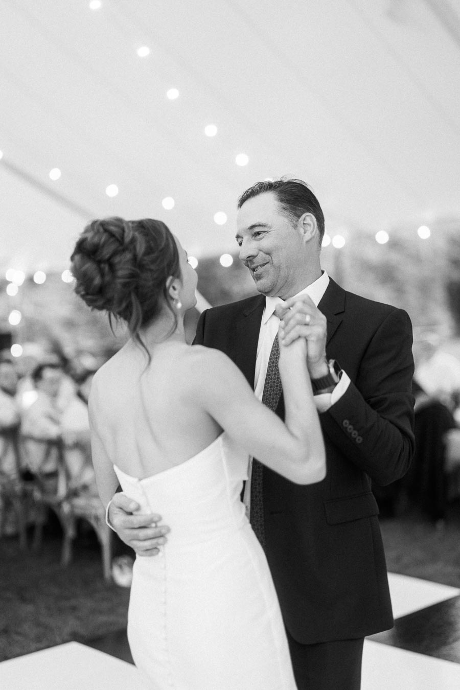 Black and white wedding photo of a bride dancing with a man inside a tent with string lights near Chatham-Kent, Ontario, Canada.