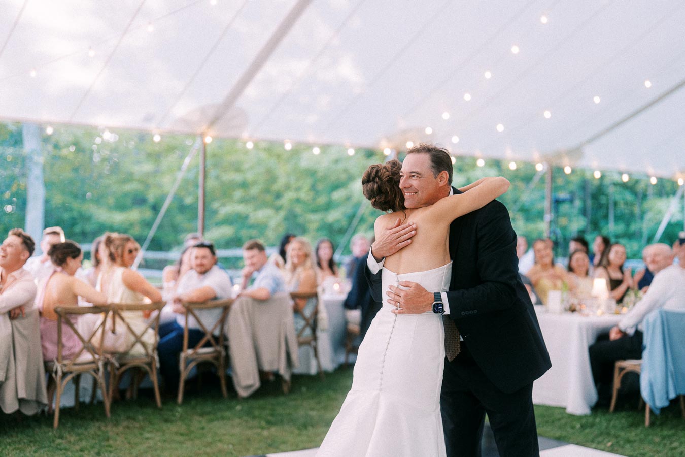 Bride hugging an older man during an outdoor tented wedding reception near Chatham-Kent, Ontario, with seated guests, tables, and string lights.