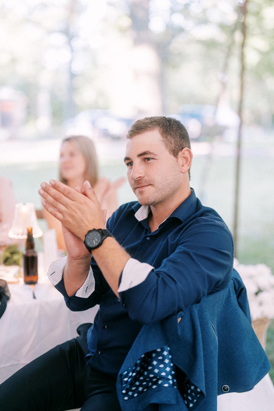 Man in a navy shirt applauding during an outdoor tented wedding dinner near Chatham-Kent, Ontario, with guests and soft garden background.
