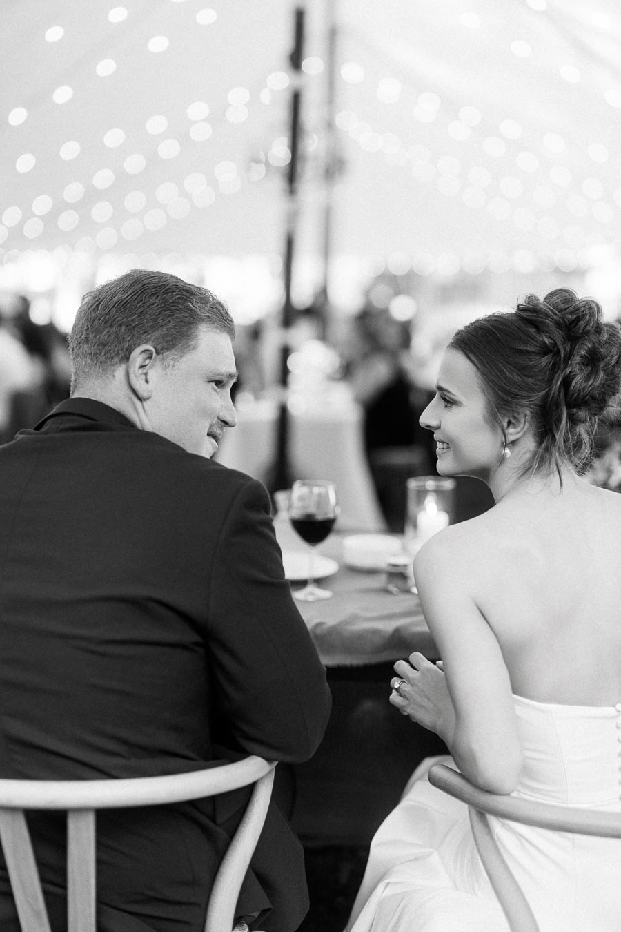 Black and white photo of a bride and groom smiling at a table during a tented wedding reception near Chatham-Kent, Ontario.