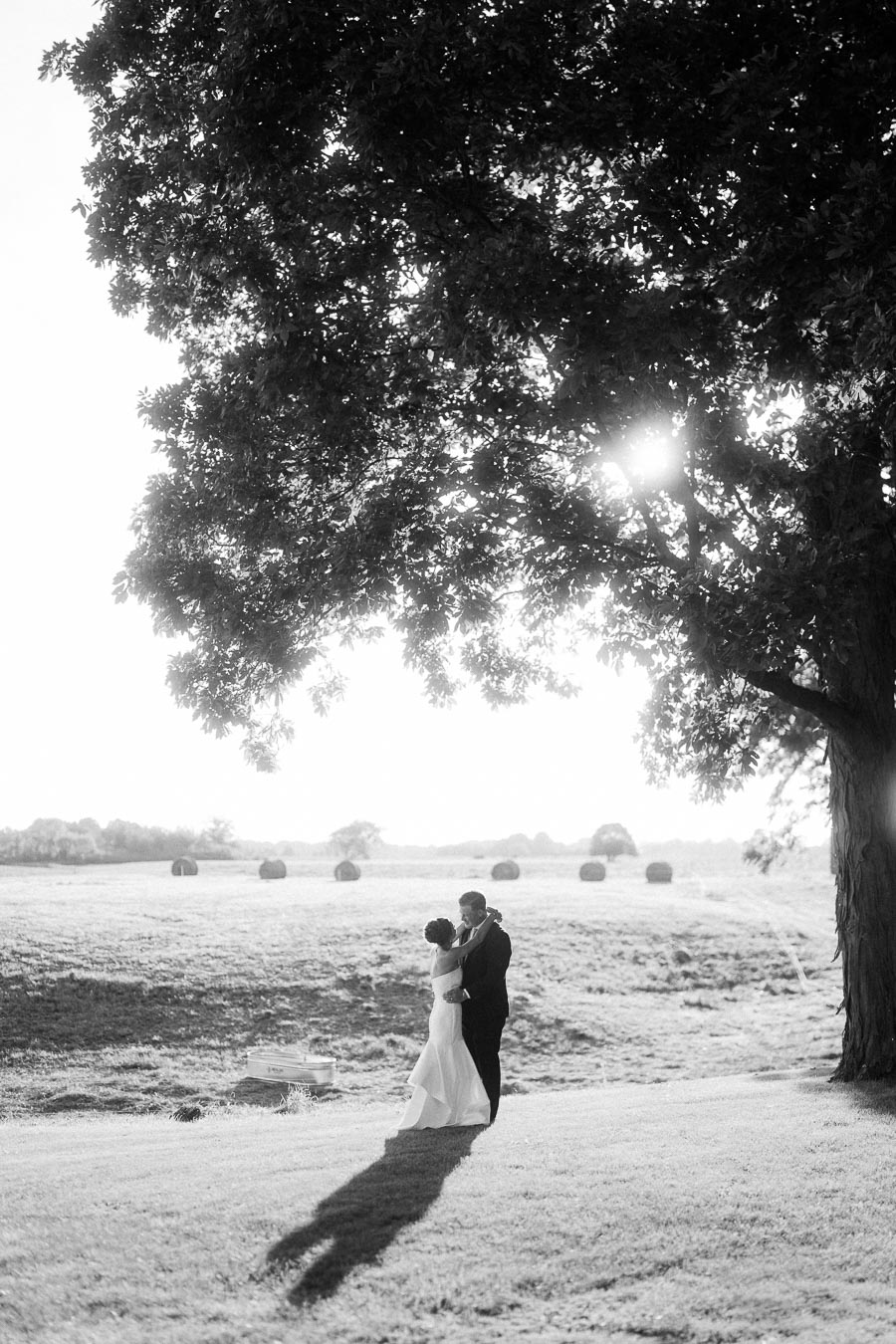 Black and white outdoor farm wedding portrait of a bride and groom embracing under a large tree near Chatham-Kent, Ontario, with hay bales in a rural field.