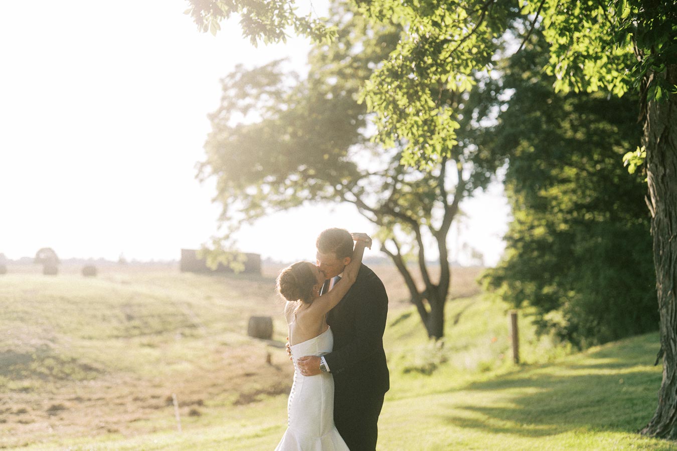 Bride and groom kissing under trees beside a farm field at a private estate wedding near Chatham-Kent, Ontario, Canada.
