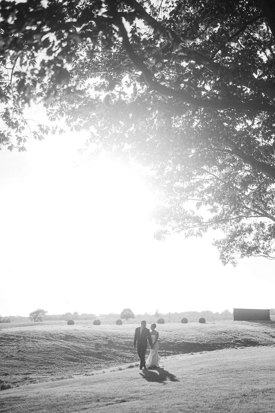 Black and white outdoor wedding portrait of a bride and groom walking across a farm field near Chatham-Kent, Ontario, Canada.