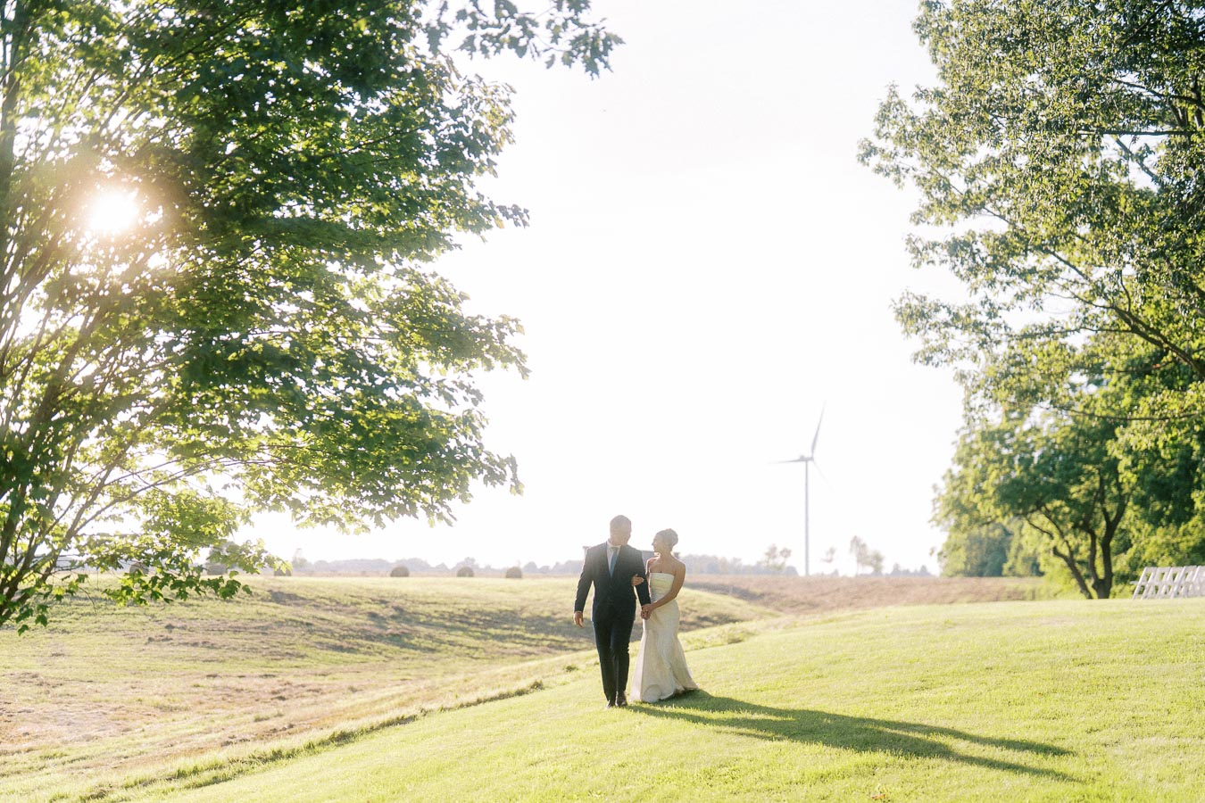 Bride and groom walking across a grassy private farm estate near Chatham-Kent, Ontario, with trees, sunset light, and a wind turbine in the background.