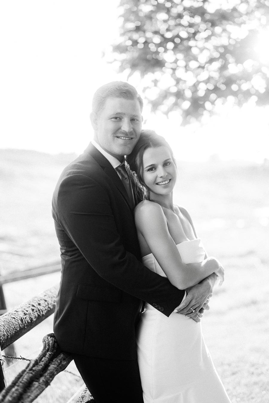 Black and white portrait of bride and groom embracing outdoors by a rustic fence at a private farm wedding near Chatham-Kent, Ontario.