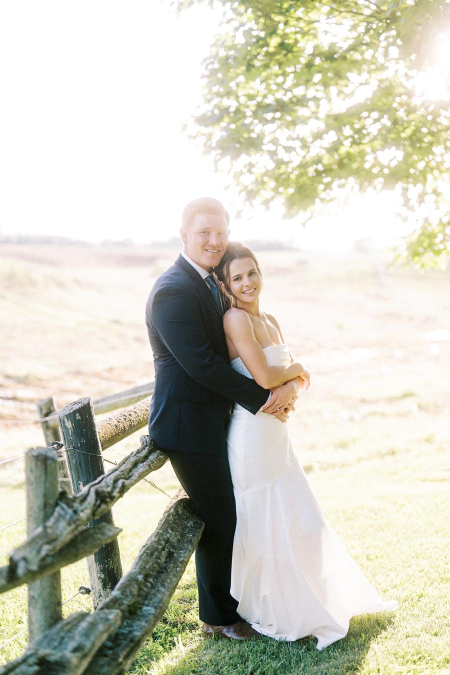 Bride and groom embracing by a rustic wooden fence on a sunny farm estate near Chatham-Kent, Ontario.