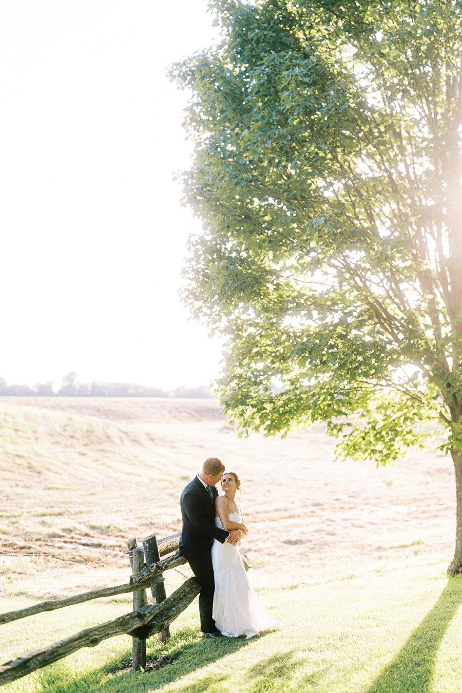 Bride and groom standing by a rustic fence under a tree on a sunlit farm field near Chatham-Kent, Ontario.