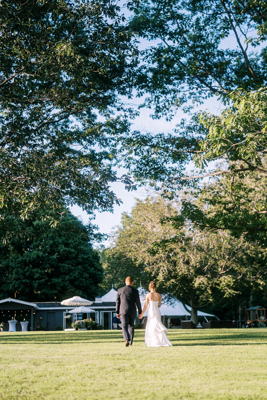 Bride and groom walking hand in hand across a grassy private estate toward a white tented farm wedding reception near Chatham-Kent, Ontario, Canada.