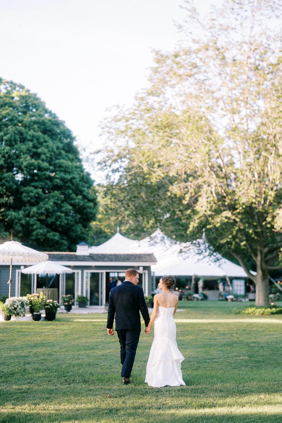 Bride and groom walking hand in hand across a lawn toward a white tented wedding reception on a private estate near Chatham-Kent, Ontario.