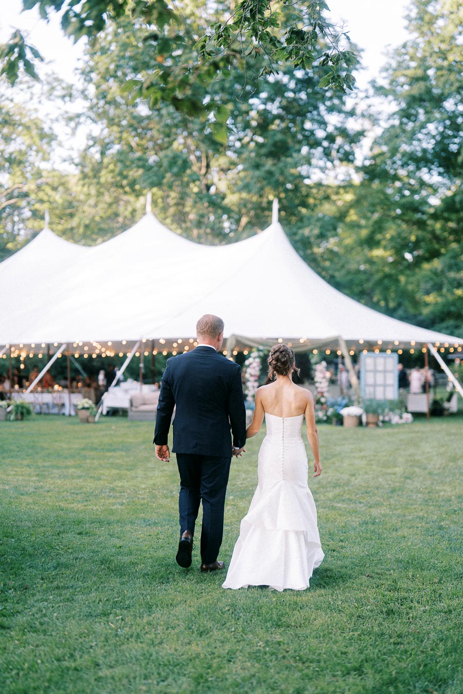 Bride and groom walking hand in hand across a lawn toward a white tented wedding reception near Chatham-Kent, Ontario, Canada.