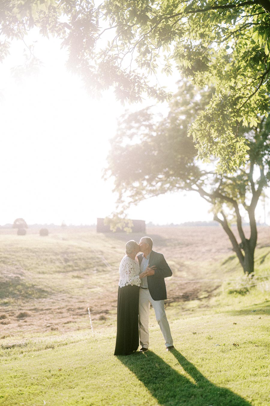 Older couple kissing outdoors on a sunlit farm lawn near Chatham-Kent, Ontario, with trees, fields, and warm evening light.