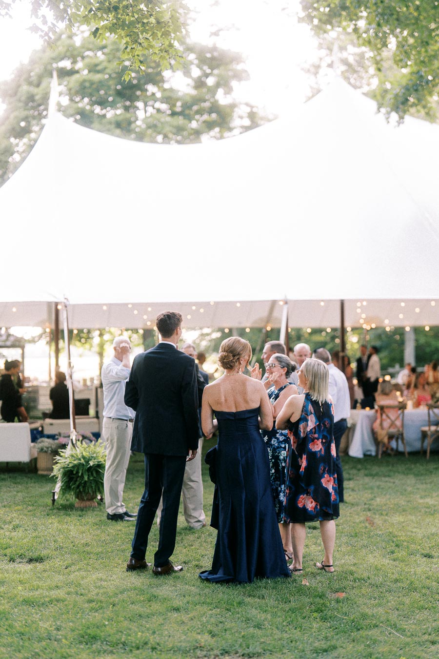 Wedding guests talking on a grassy private estate lawn beside a white reception tent with string lights near Chatham-Kent, Ontario, Canada.