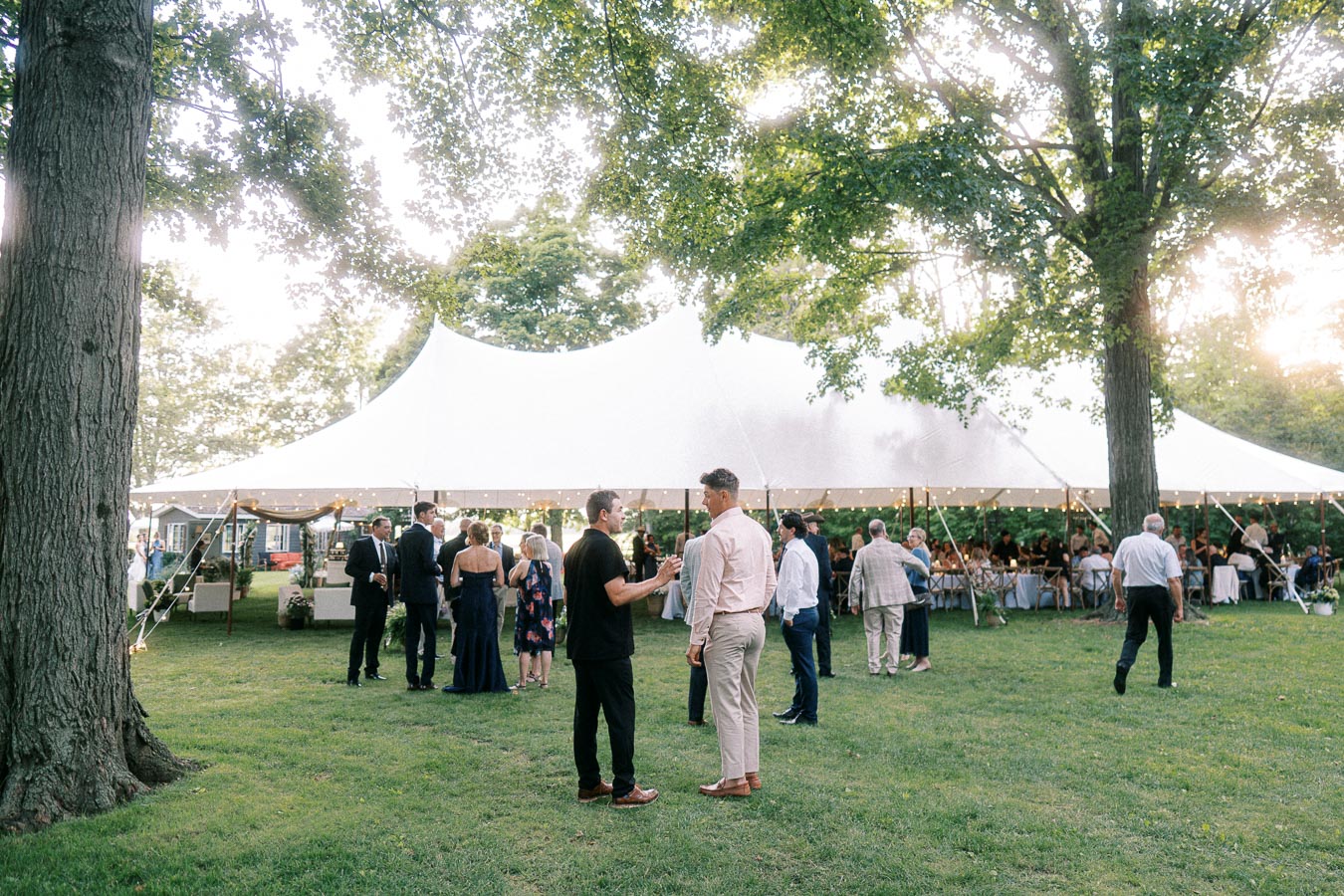 Outdoor tented wedding reception on a private farm estate near Chatham Kent, Ontario, with guests mingling under large trees at sunset.