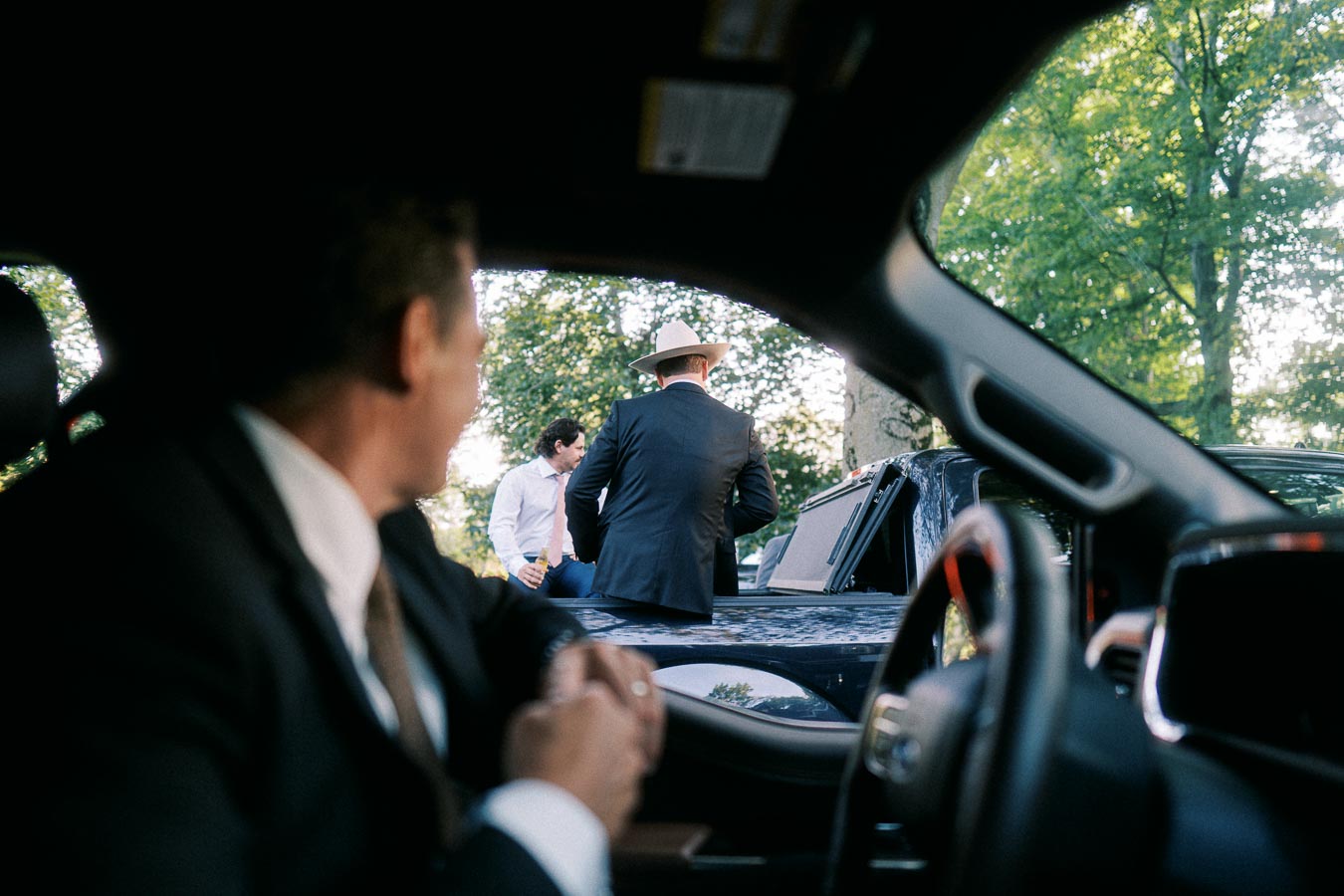 Groomsman seated inside a vehicle watching men in suits beside a pickup truck during an outdoor farm wedding near Chatham-Kent, Ontario.