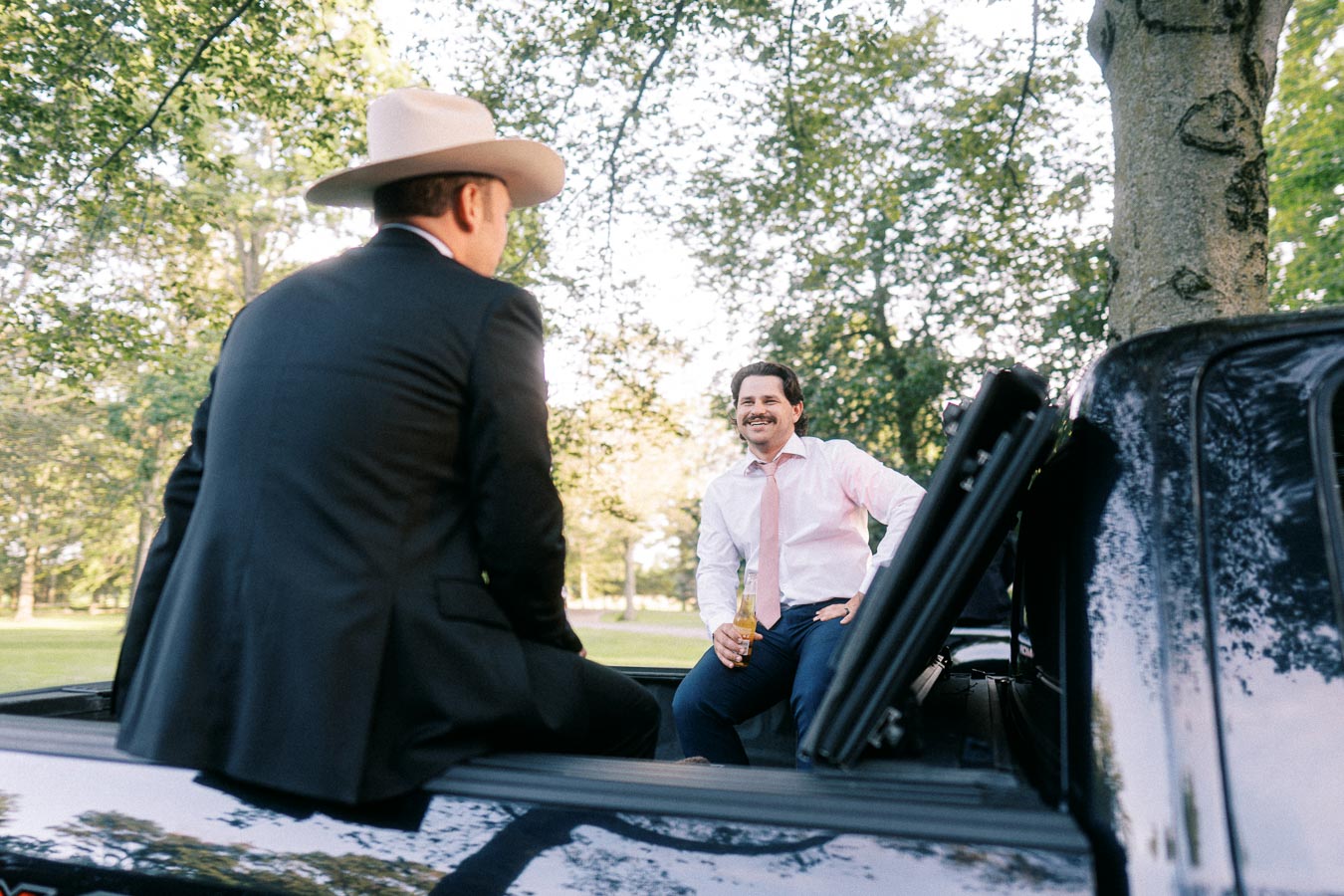 Two men in wedding attire sit on the back of a pickup truck under trees during an outdoor farm wedding near Chatham-Kent, Ontario.