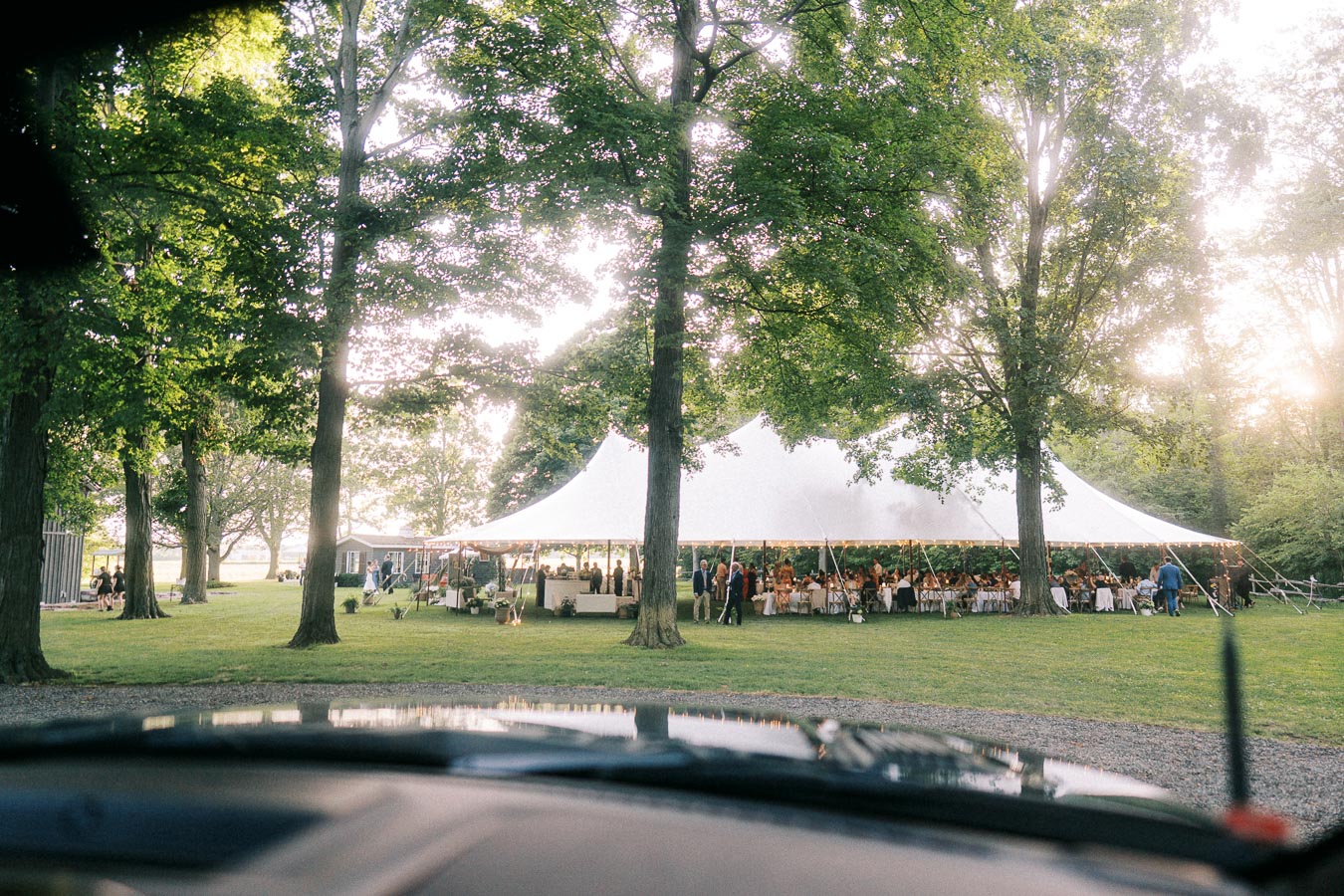 Outdoor tented wedding dinner on a grassy estate with guests seated under a white canopy among tall trees near Chatham-Kent, Ontario, Canada.