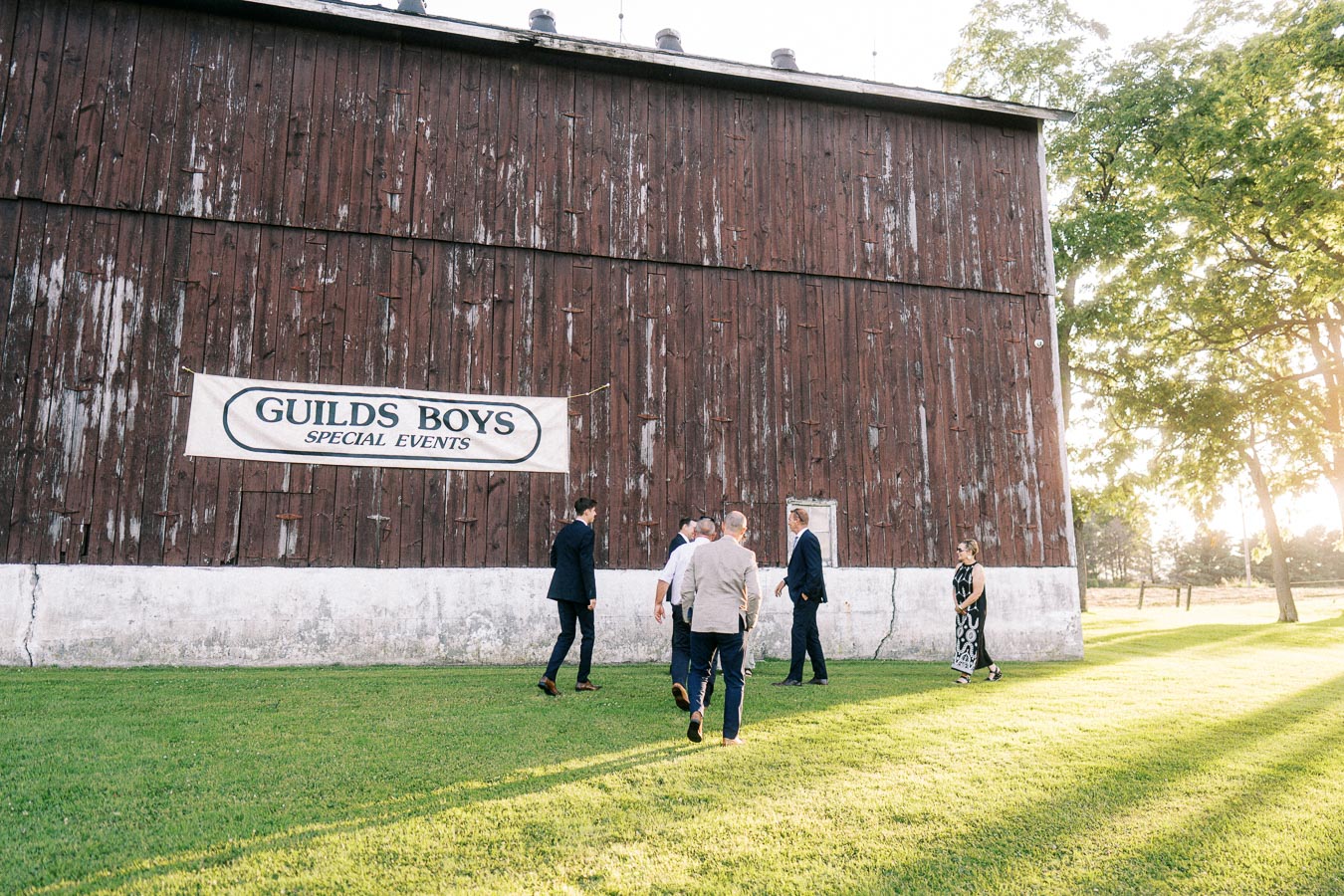 Wedding guests walk across a grassy private farm estate beside a weathered red barn with Guilds Boys Special Events sign near Chatham Kent, Ontario.