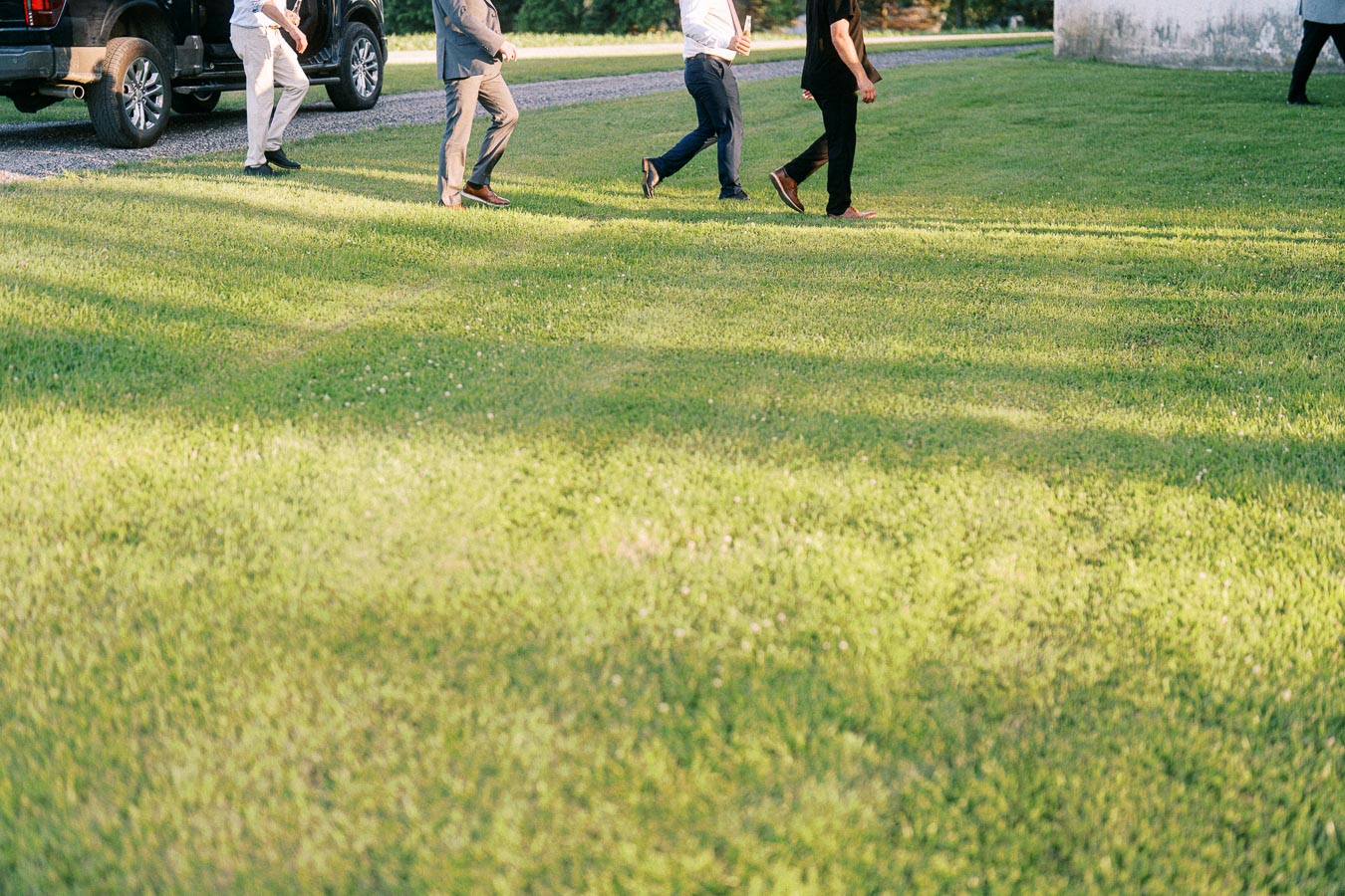 Wedding guests walking across a sunny farm lawn near parked vehicles on a private property in Chatham-Kent, Ontario, Canada.